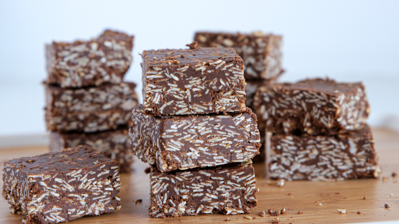 Several pieces of chocolate coconut squares are neatly stacked on a wooden surface. The squares have a rich, dark chocolate color with visible shredded coconut pieces mixed in. The background is simple and out of focus, emphasizing the texture of the treats.