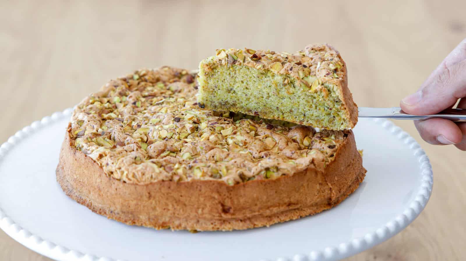 A hand lifts a slice from a pistachio cake on a white cake stand. The cake features a golden crust and is topped with chopped pistachios, revealing a green, moist interior. The background is a light wood surface.