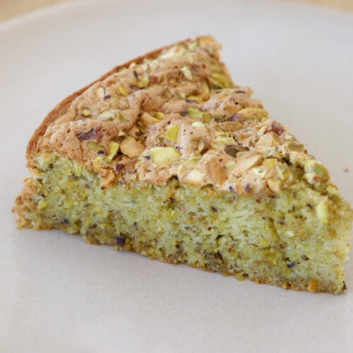 A slice of flourless pistachio cake sits on a plain white plate. The cake appears moist with a greenish hue and is topped with chopped pistachios. The plate rests on a light wooden surface.