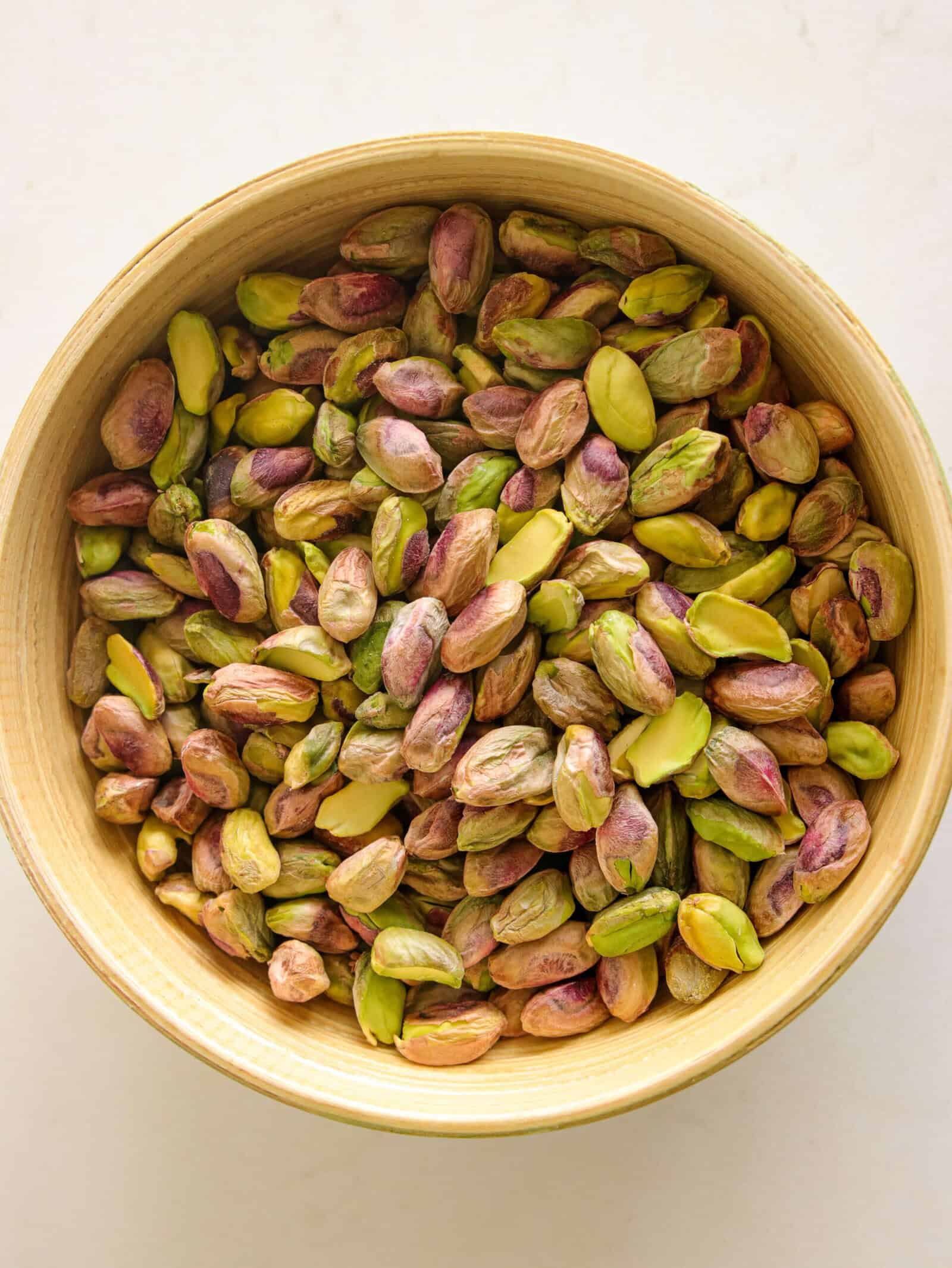 A wooden bowl filled with unshelled pistachios, showing a mix of green and purple hues, placed on a light-colored surface.