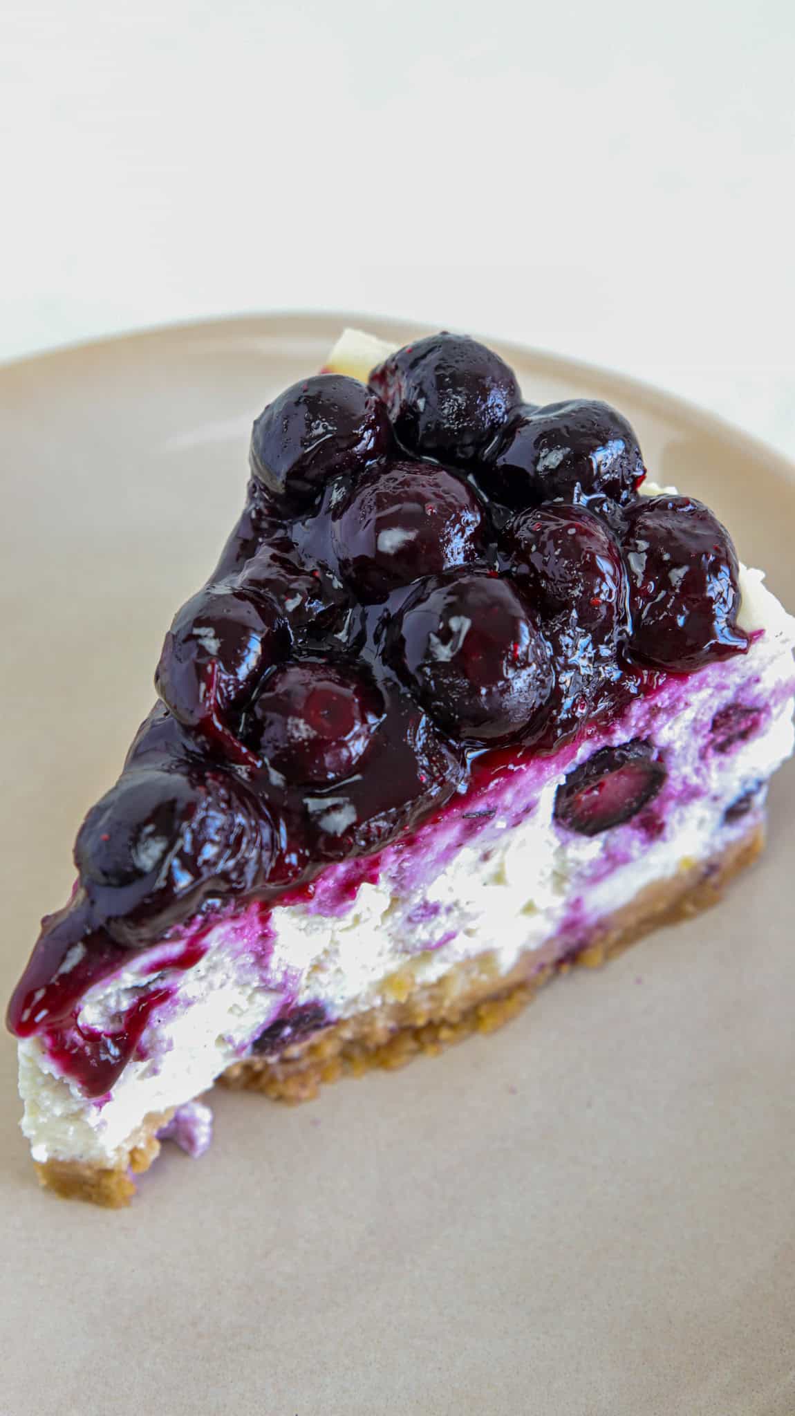 A slice of cheesecake topped with glossy blueberry compote sits on a beige plate, showing layers of creamy filling, blueberry swirls, and a crumbly crust.