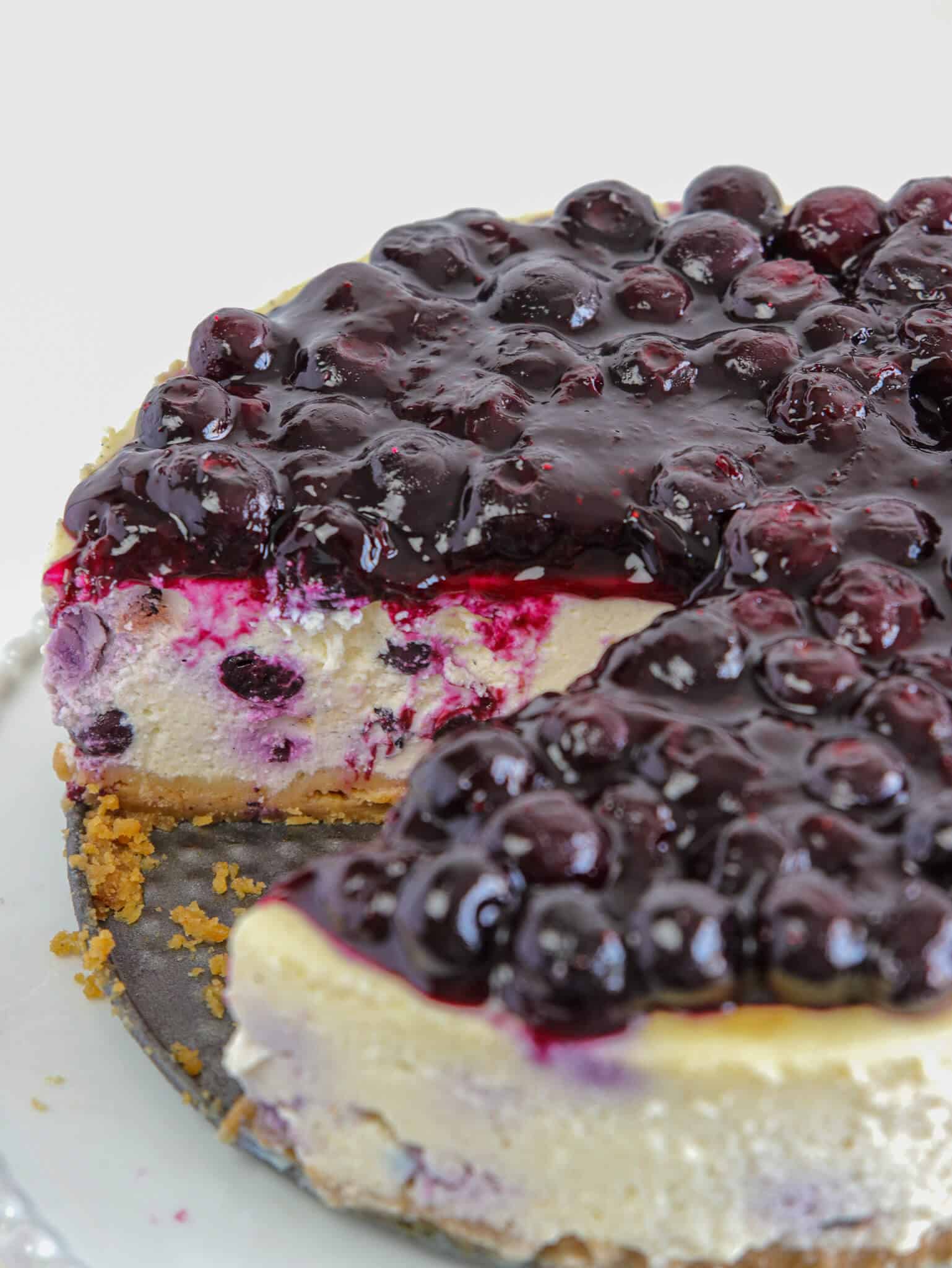A cheesecake topped with glossy blueberry compote sits on a plate, with a large slice removed to reveal the creamy interior and graham cracker crust.