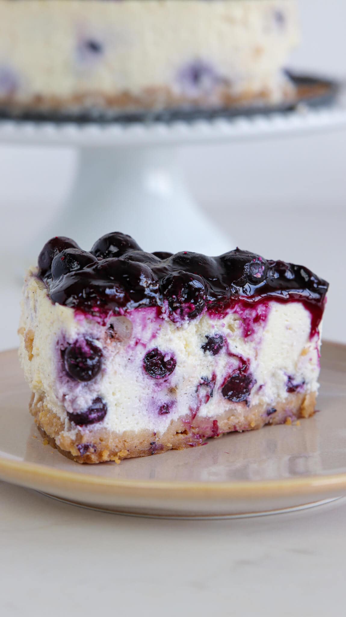 A slice of blueberry cheesecake with a crumbly crust and blueberry topping sits on a beige plate, with the rest of the cheesecake on a cake stand in the background.