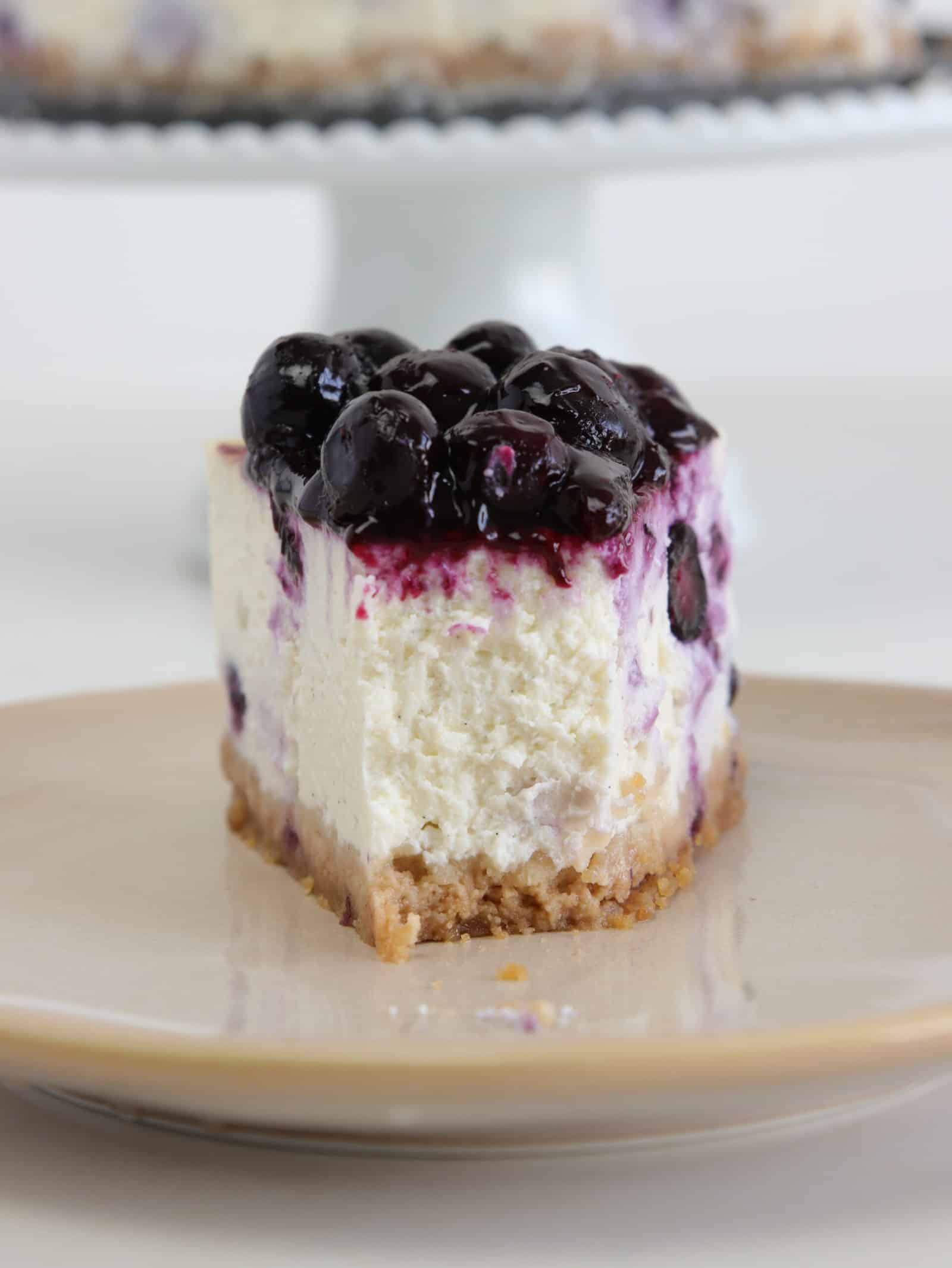 A slice of cheesecake with a crumbly crust and topped with glossy blueberry sauce sits on a beige plate, with the rest of the cheesecake blurred in the background.