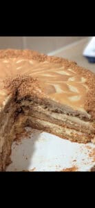 A multi-layered cake with creamy frosting and a caramel glaze on top, partially sliced to reveal the inner layers. Crumbled cake or cookie pieces decorate the edges.