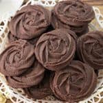 A plate of chocolate cookies arranged on a decorative white doily. The cookies are shaped like roses and placed on a wooden surface with a gold fork and a white cup in the background.