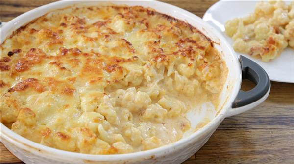 A baked cauliflower cheese dish in a white baking dish with handles. The dish has a browned, cheesy crust on top and a portion has been scooped out and served onto a nearby white plate. The creamy, cheesy sauce is visible where the portion was removed.