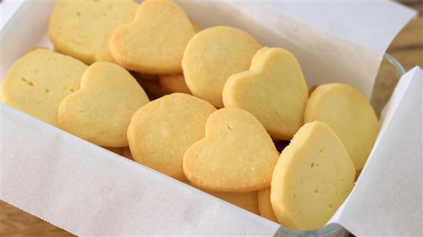 A glass dish lined with white parchment paper holds a batch of freshly baked cookies. The cookies are a light golden color and are shaped like hearts and circles. They appear to be evenly baked with a smooth texture.