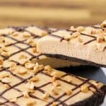 A slice of peanut butter pie is being lifted from the whole pie. The pie has a chocolate crust, creamy peanut butter filling, and is topped with a lattice pattern of chocolate drizzle and scattered chopped peanuts. The background consists of a wooden surface.