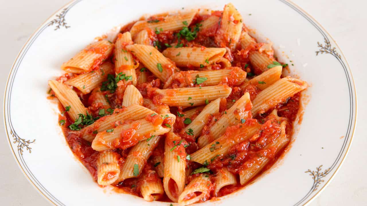 A white plate contains a serving of penne pasta topped with a rich tomato sauce and garnished with finely chopped parsley. The sauce is thick and chunky, covering the pasta evenly. The dish is placed on a light-colored surface.