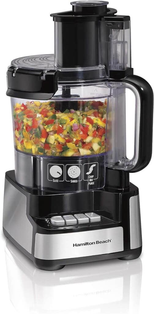 A Hamilton Beach food processor filled with chopped vegetables sits on a reflective surface. The machine has a transparent container, black base with control buttons for slice, shred, and chop/puree functions, and a black handle on the right.