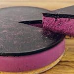 A vibrant purple cheesecake with a glossy, dark purple topping sits on a wooden surface. One slice has been cut and is being lifted, revealing the rich, creamy texture of the filling and the light brown crust.