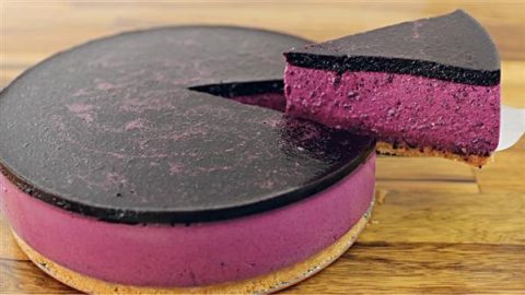 A vibrant purple cheesecake with a glossy, dark purple topping sits on a wooden surface. One slice has been cut and is being lifted, revealing the rich, creamy texture of the filling and the light brown crust.