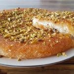 A round, golden-brown dessert served on a white plate, topped with chopped pistachios. A triangular slice has been cut and is being lifted, revealing a layer of cheese or cream inside. The dessert is placed on a wooden surface.