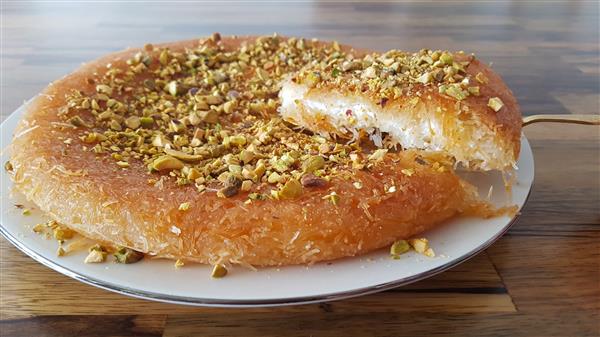 A round, golden-brown dessert served on a white plate, topped with chopped pistachios. A triangular slice has been cut and is being lifted, revealing a layer of cheese or cream inside. The dessert is placed on a wooden surface.