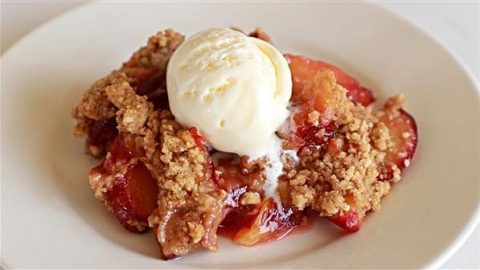 A close-up of a plate with a serving of fruit crumble topped with a scoop of vanilla ice cream. The dessert features baked fruit pieces and a golden-brown crumbly topping. The ice cream is slightly melting over the warm crumble.