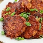 A white plate filled with crispy, golden-brown potato latkes garnished with chopped green onions, set on a wooden table. The latkes are stacked slightly on top of each other.