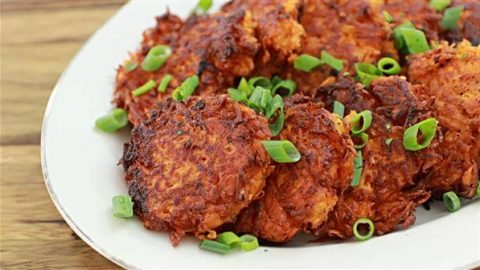 A white plate filled with crispy, golden-brown potato latkes garnished with chopped green onions, set on a wooden table. The latkes are stacked slightly on top of each other.