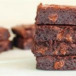 A stack of four gooey, chocolate brownies is pictured in the foreground, showcasing melted chocolate chips inside. In the background, more brownies are slightly blurred on a light-colored surface. The brownies have a rich, dense texture.