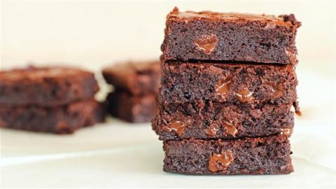 A stack of four gooey, chocolate brownies is pictured in the foreground, showcasing melted chocolate chips inside. In the background, more brownies are slightly blurred on a light-colored surface. The brownies have a rich, dense texture.