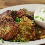 A plate of golden-brown vegetable fritters garnished with chopped green onions, accompanied by a small bowl of white dipping sauce topped with more green onions. The dish is set on a wooden table.