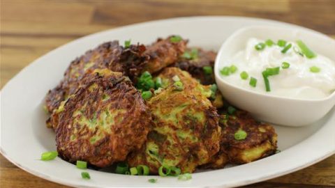 A plate of golden-brown vegetable fritters garnished with chopped green onions, accompanied by a small bowl of white dipping sauce topped with more green onions. The dish is set on a wooden table.