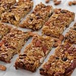 A cluster of homemade granola bars arranged neatly on a baking sheet, surrounded by scattered almonds. Each bar is packed with oats, nuts, and dried fruits, showcasing a crunchy and wholesome texture. The baked bars are lightly golden and ready to eat.