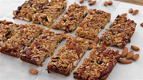 A cluster of homemade granola bars arranged neatly on a baking sheet, surrounded by scattered almonds. Each bar is packed with oats, nuts, and dried fruits, showcasing a crunchy and wholesome texture. The baked bars are lightly golden and ready to eat.