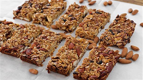 A cluster of homemade granola bars arranged neatly on a baking sheet, surrounded by scattered almonds. Each bar is packed with oats, nuts, and dried fruits, showcasing a crunchy and wholesome texture. The baked bars are lightly golden and ready to eat.