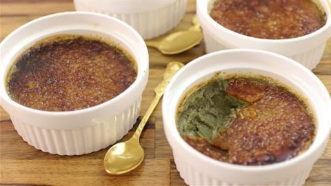 Four white ramekins, each filled with crème brûlée, are placed on a wooden surface. Two small golden spoons lie beside them. One of the ramekins has a portion of its caramelized sugar top cracked open, revealing the creamy custard inside.