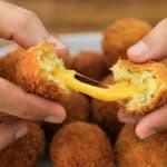 Close-up of two hands pulling apart a fried cheese ball, revealing melted cheese stretching between the halves. More fried cheese balls are seen on a plate in the background.