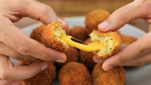 Close-up of two hands pulling apart a fried cheese ball, revealing melted cheese stretching between the halves. More fried cheese balls are seen on a plate in the background.