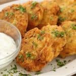 A plate of golden-brown fritters garnished with chopped herbs is served alongside a clear bowl filled with a white creamy dipping sauce. The fritters appear to be homemade and freshly cooked, with a crisp texture visible on their surface.