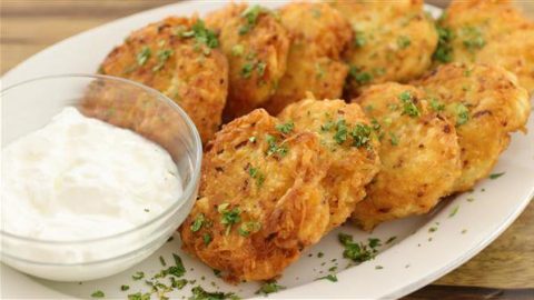 A plate of golden-brown fritters garnished with chopped herbs is served alongside a clear bowl filled with a white creamy dipping sauce. The fritters appear to be homemade and freshly cooked, with a crisp texture visible on their surface.