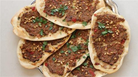 A plate of Middle Eastern flatbreads topped with seasoned minced meat, pine nuts, and fresh parsley. The flatbreads are baked to a golden-brown perfection and presented on a metallic serving tray.