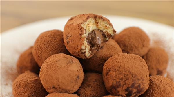 Close-up of several cocoa-dusted chocolate truffles piled on a plate. One truffle at the top has a bite taken out, revealing a creamy chocolate filling. The background is blurred, drawing focus to the truffles.