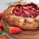 A rustic strawberry galette rests on a wooden board. Surrounding the galette are several fresh strawberries. In the background, silver forks are visible on a light-colored surface. The galette features a golden-brown crust with slices of strawberry filling.