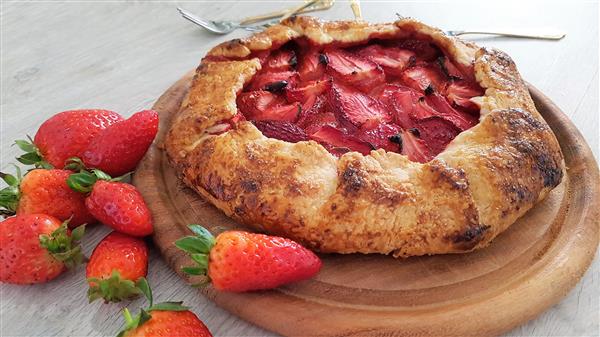 A rustic strawberry galette rests on a wooden board. Surrounding the galette are several fresh strawberries. In the background, silver forks are visible on a light-colored surface. The galette features a golden-brown crust with slices of strawberry filling.