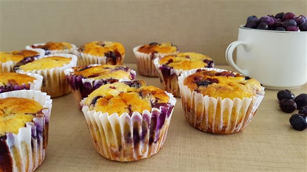 Several freshly baked blueberry muffins in white paper liners are arranged on a wooden surface. A white cup filled with blueberries is placed in the background. Some blueberries have spilled onto the surface next to the cup.