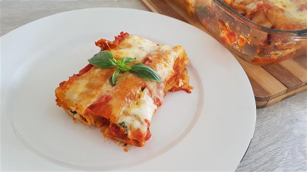 A white plate holds a serving of cheesy lasagna garnished with fresh basil leaves. In the background, a glass baking dish with more lasagna sits on a wooden cutting board. The lasagna is layered with tomato sauce, noodles, and melted cheese.