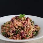 A bowl of bulgur salad on a white plate. The salad is mixed with finely chopped parsley, tomatoes, onions, and cubes of beets, garnished with a mint leaf on top. The background is a simple, dark, plain color, enhancing the vivid colors of the salad.