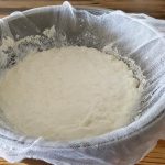 A stainless steel colander lined with cheesecloth is filled with freshly made, soft-looking cheese. The colander is placed on a wooden surface. The cheesecloth is draped over the edges of the colander, with some parts slightly hanging over.