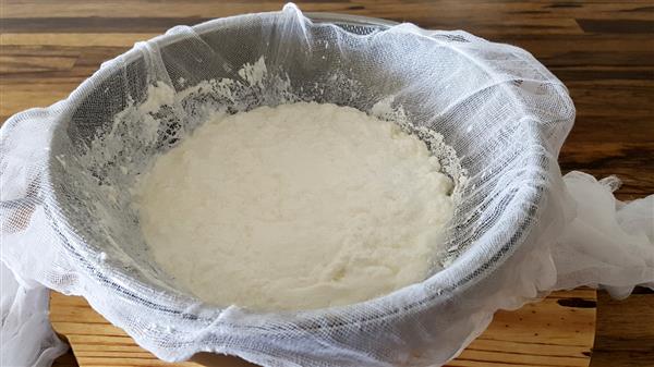 A stainless steel colander lined with cheesecloth is filled with freshly made, soft-looking cheese. The colander is placed on a wooden surface. The cheesecloth is draped over the edges of the colander, with some parts slightly hanging over.