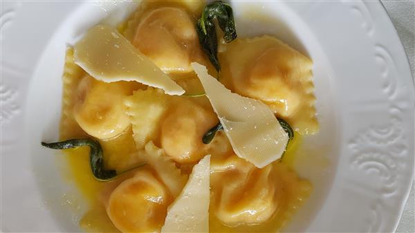 A white plate holds a serving of ravioli topped with melted butter and garnished with fresh sage leaves and several thin shavings of Parmesan cheese. The edges of the plate are decorated with an intricate embossed pattern.