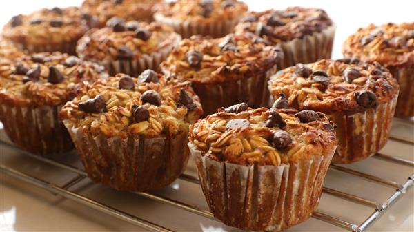 Close-up of baked oatmeal muffins with chocolate chips on top, lined in white paper cups. The muffins are cooling on a metal rack, showcasing their golden-brown, slightly textured surface with oats and chocolate chips clearly visible.