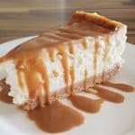 A slice of creamy cheesecake with a graham cracker crust sits on a white plate. Caramel sauce is generously drizzled over the top, running down the sides and pooling on the plate. The background features a wooden table surface.