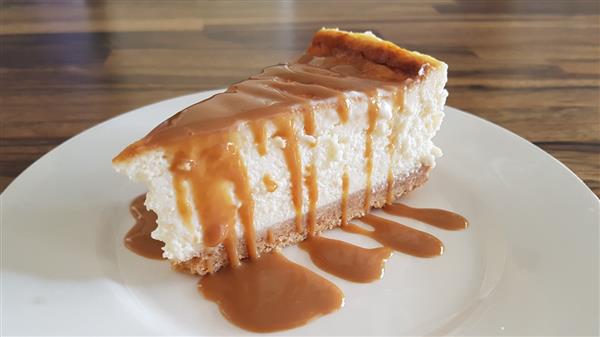 A slice of creamy cheesecake with a graham cracker crust sits on a white plate. Caramel sauce is generously drizzled over the top, running down the sides and pooling on the plate. The background features a wooden table surface.