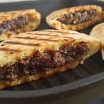 Four grilled, partially sliced pita breads filled with a savory mixture of ground meat, herbs, and spices. The pitas are browned with grill marks, and the filling is visible, showcasing a juicy, well-seasoned meat blend. They are served on a black grill pan.