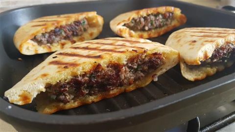 Four grilled, partially sliced pita breads filled with a savory mixture of ground meat, herbs, and spices. The pitas are browned with grill marks, and the filling is visible, showcasing a juicy, well-seasoned meat blend. They are served on a black grill pan.