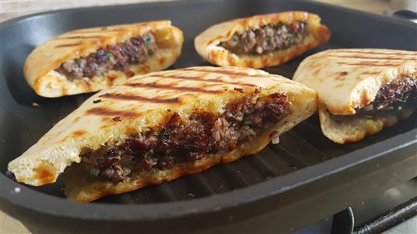 Four grilled, partially sliced pita breads filled with a savory mixture of ground meat, herbs, and spices. The pitas are browned with grill marks, and the filling is visible, showcasing a juicy, well-seasoned meat blend. They are served on a black grill pan.